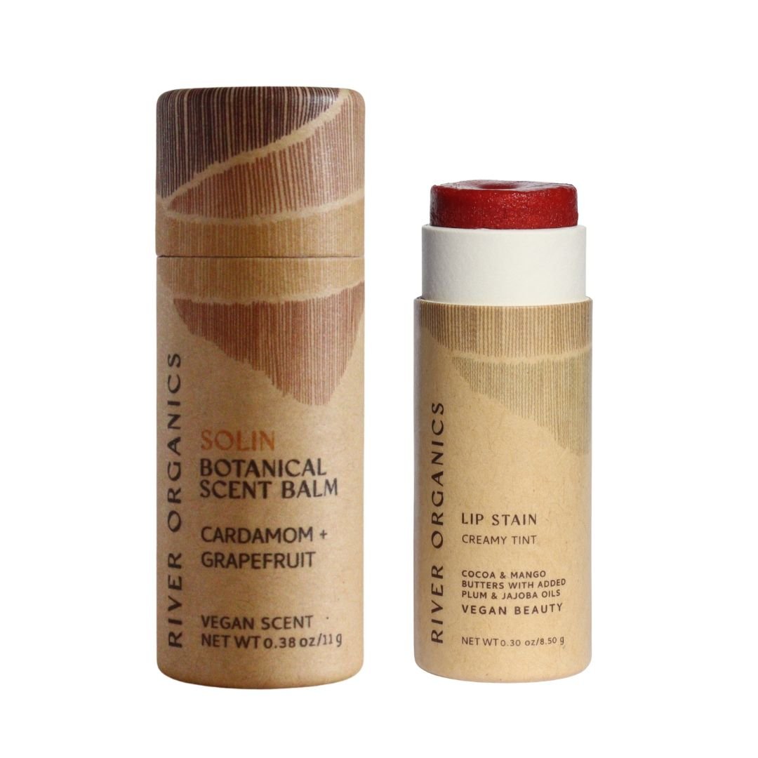 River Organics’ Lip + Scent Balm Bundle features two cylindrical products on a white background: a closed Botanical Scent Balm—Cardamom + Grapefruit, and an open Vegan Lip Balm showcasing its creamy red tint.