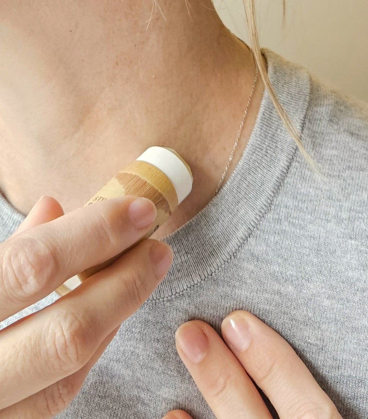 A person applies River Organics' Still Earth: Vetiver + Sandalwood Botanical Scent Balm to the side of their neck while wearing a light gray shirt and a thin silver necklace. Only their hand, neck, and part of their face are visible.