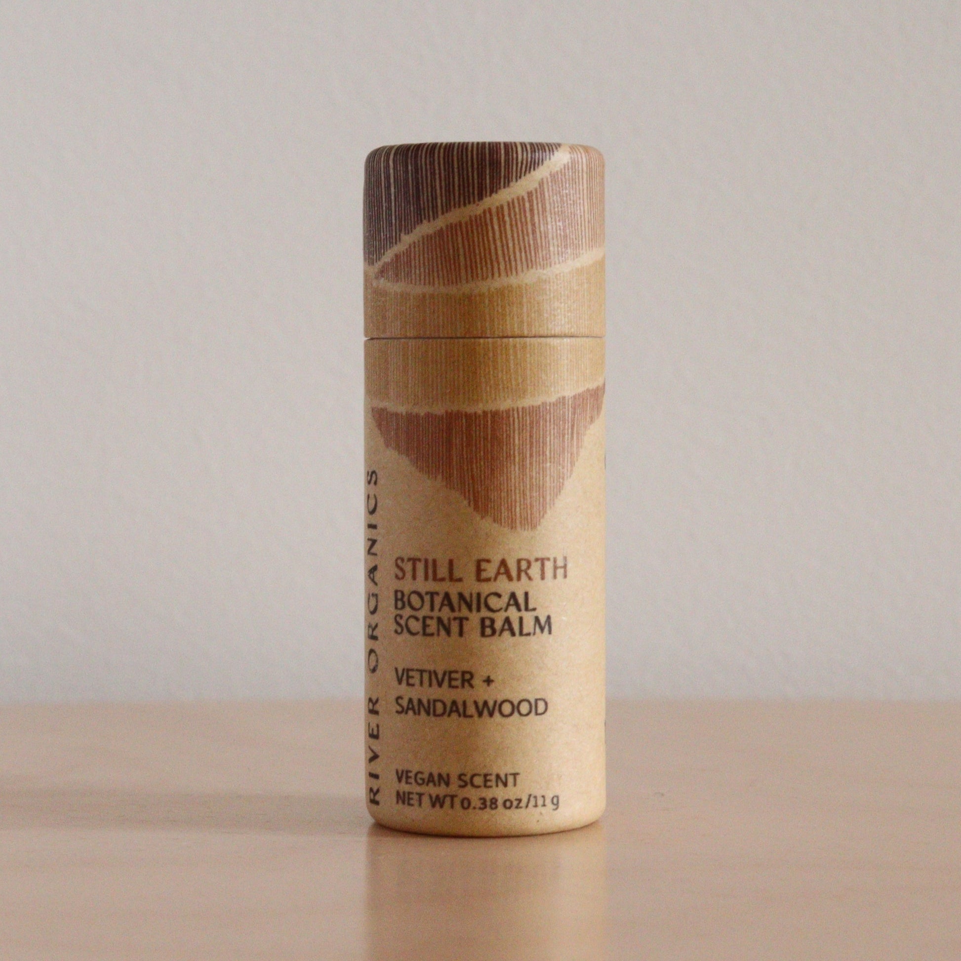 A cylindrical cardboard container of River Organics’ Still Earth: Vetiver + Sandalwood - Botanical Scent Balm stands upright on a plain surface. This botanical balm also serves as a natural perfume.