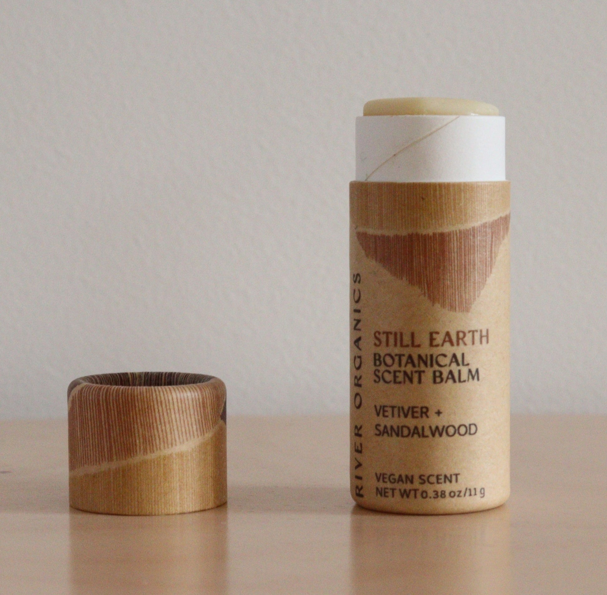 A cylindrical cardboard tube of River Organics Still Earth: Vetiver + Sandalwood Botanical Scent Balm with the cap removed and set beside it, exposing the natural perfume balm inside.