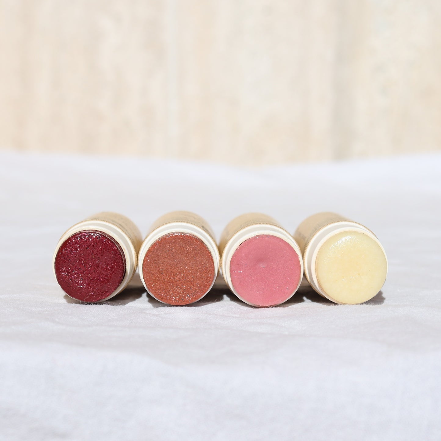 Four River Organics Lip + Concealer Set tubes are lying horizontally on a white surface, their ends facing the camera. The balms display a variety of shades—deep red, bronze, pink, and cream—offering vibrant color options for your clean beauty routine. The background is softly blurred.