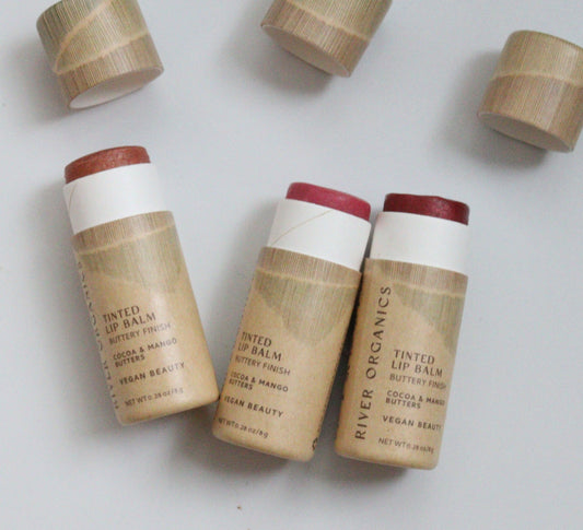 Three tubes of River Organics Tinted Vegan Lip Balm – Plastic-Free, Organic & Hydrating—with caps off—show pink and red shades. The beige, eco-friendly packaging lists ingredients like cocoa butter and highlights its vegan formula.