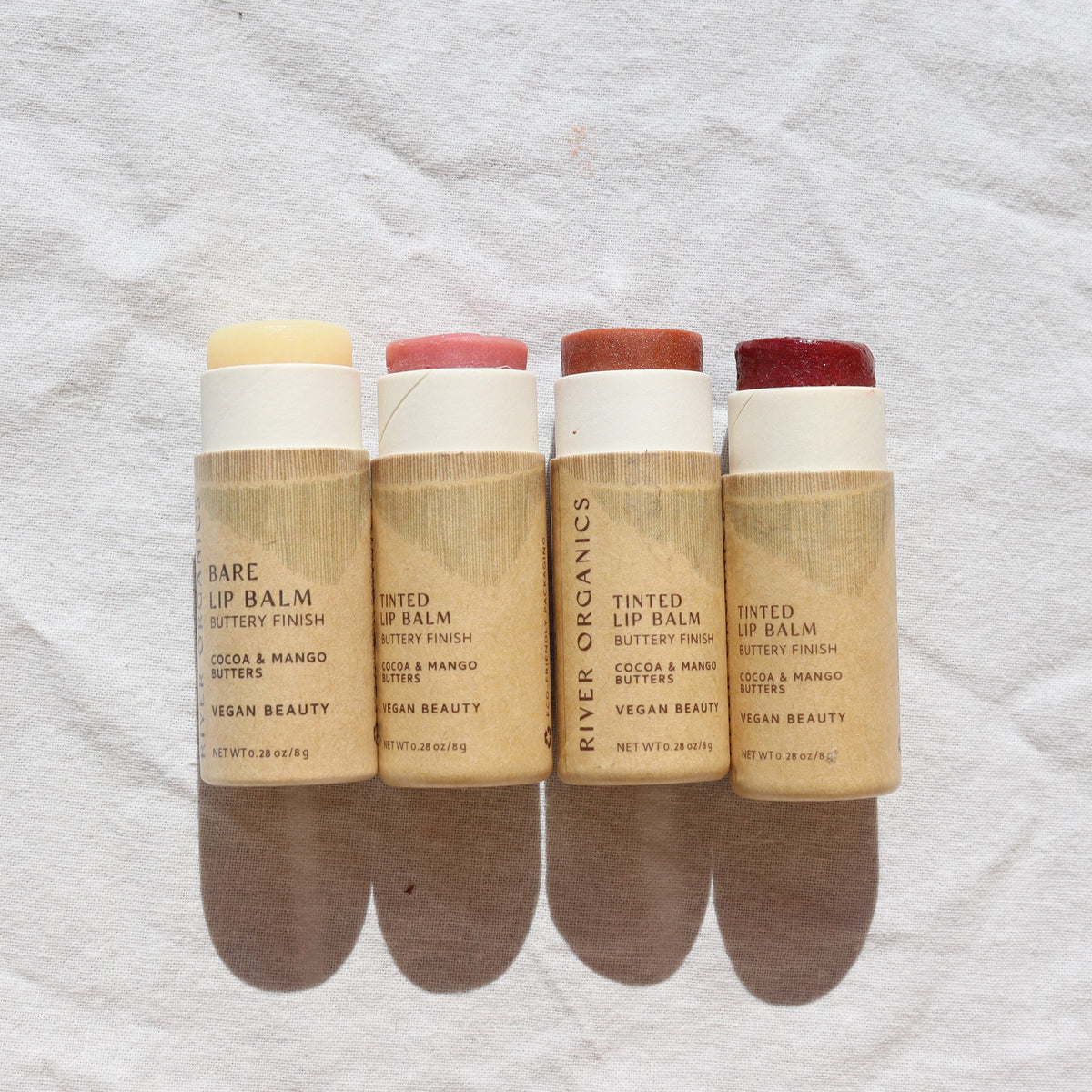 Four tubes of River Organics Imperfect Lip Balms and Lip Stains – New Full Size Products, each featuring a unique tint, are showcased in plastic-free packaging that stands upright on a light surface, displaying their beautiful shades: beige, pink, peach, and deep red.