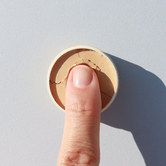 A finger presses into a round container of River Organics Zero-Waste Vegan Concealer (Titanium Dioxide-Free) on a light background, casting a shadow to the right.