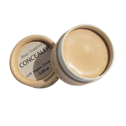 A small, round, open beige container with a beige concealer inside. The label on the cap reads, "River Organics Zero-Waste Concealer, Soft Vegan Cover, 0.26 oz." Made with Certified Organic oils and a plant-based formula, the container and cap feature a minimalist design aligned with zero waste principles.