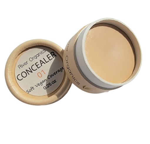 A small, round container of River Organics Imperfect Natural Hydrating Vegan Concealer with the lid off, revealing light beige cream. Made with organic plant oils for zero-waste; label: "CONCEALER 01, Soft Vegan Coverage, 0.25 oz.
