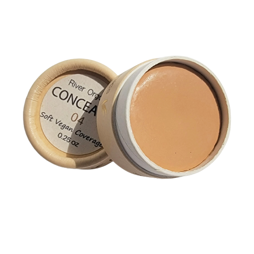 A small round container of Imperfect Natural Hydrating Vegan Concealer by River Organics, lid off and placed beside it. This zero-waste concealer offers soft, vegan coverage in a medium beige shade, made with organic plant oils.