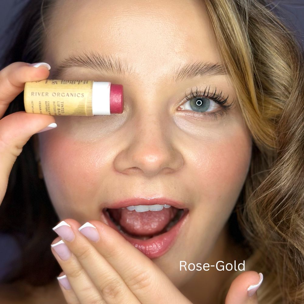 A delighted woman holds a River Organics Tinted Vegan Lip Balm near her eye. The rose-gold shade beautifully complements her light makeup and manicured nails, adding a touch of elegance with its natural ingredients.