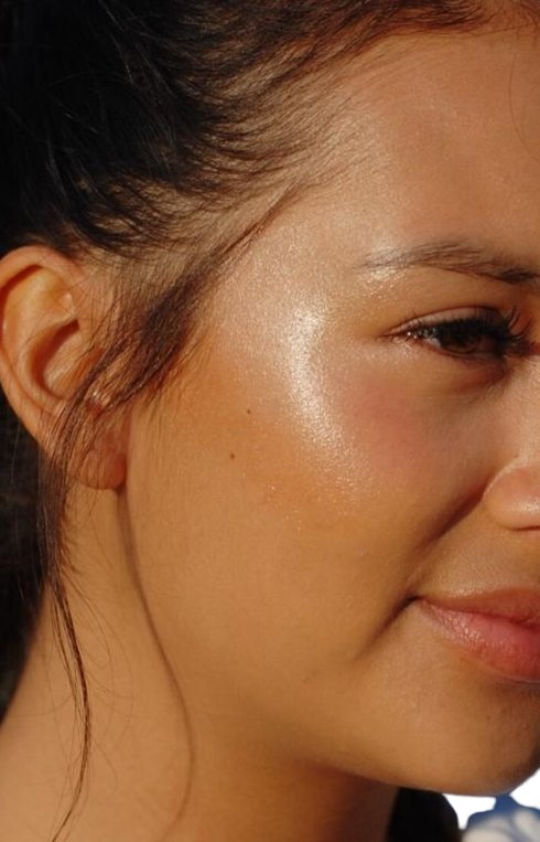 Close-up of a person's face, focusing on the cheek enhanced with River Organics' Rose Quartz Vegan Highlighter Stick. The skin appears smooth, with a natural, radiant glow. The person has dark hair pulled back and is slightly smiling.