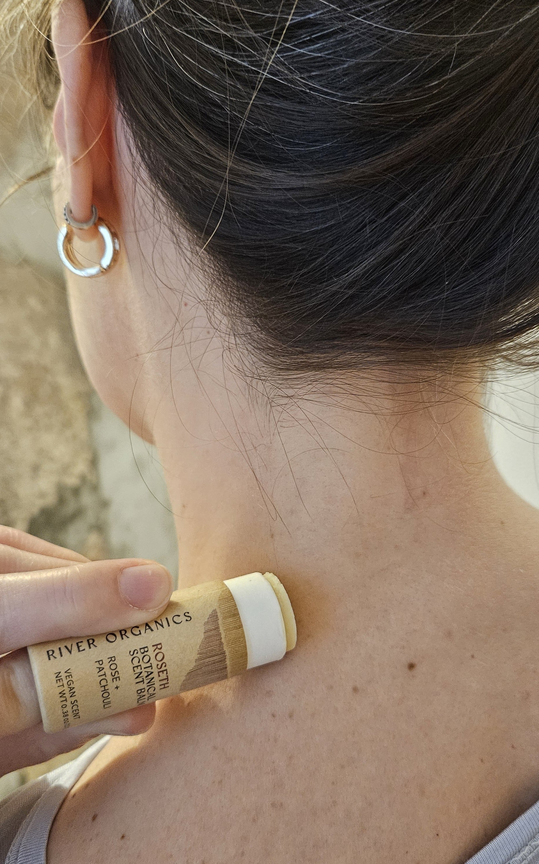 A person with brown hair in a bun applies River Organics Roseth: Rose + Patchouli - Botanical Scent Balm to the back of their neck, adding natural fragrance to their routine while wearing a tank top and silver hoop earring.