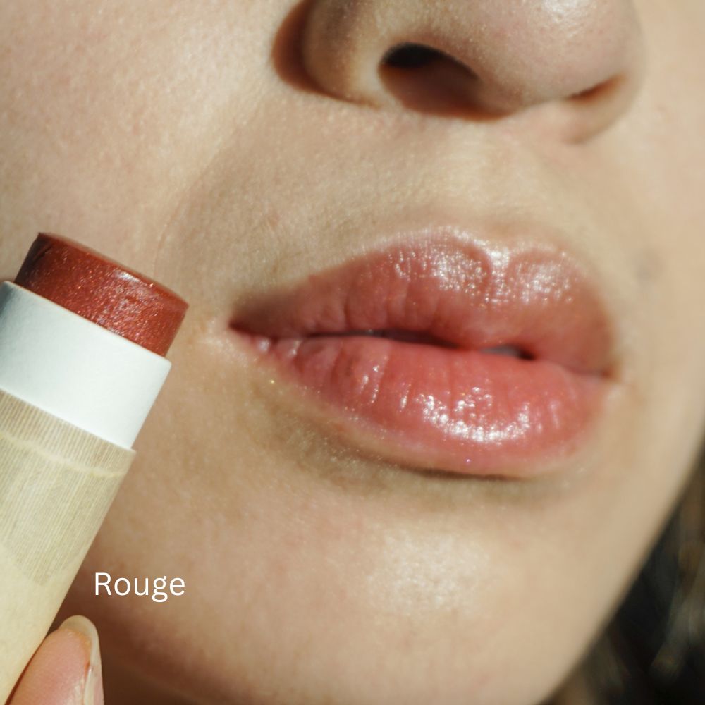 Close-up of lips with a natural, glossy finish holding River Organics' Imperfect Lip Balm and Lip Stain (new full size) in eco-friendly packaging labeled "Rouge," near the mouth.