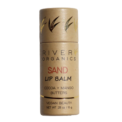 The image shows a cylindrical tube of River Organics Tinted Vegan Lip Balm | Sand. The eco-friendly packaging is a light brown color with gold accents and text. Labeled as being made with cocoa and mango butters, this vegan lip balm is also noted to be natural tinted. The net weight is 0.28 oz or 8 g.