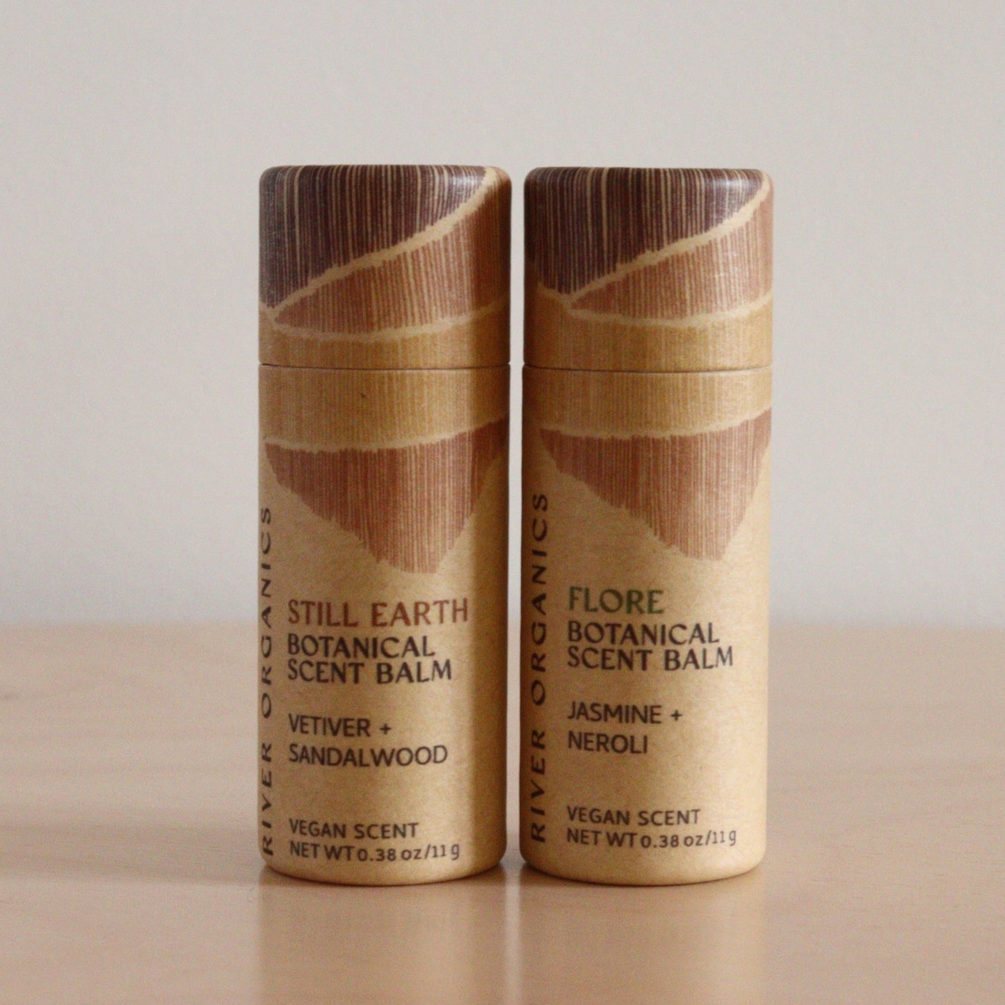 Two cylindrical River Organics Botanical Scent Balms from the Set of 2 stand on a light surface. Labeled "Still Earth" and "Flore," these solid perfumes feature natural designs and are enriched with plant oils.
