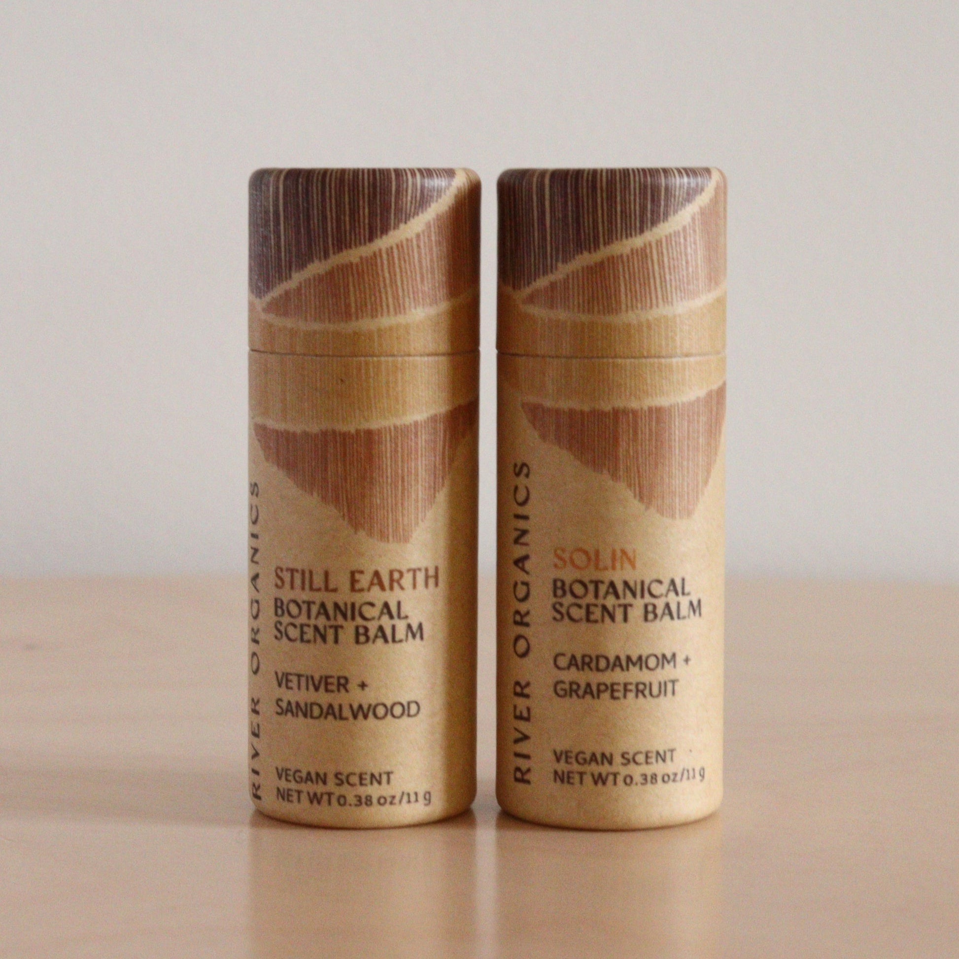 Two River Organics Botanical Scent Balms from the "Set of 2, 3 or 4"—"Still Earth" (Vetiver + Sandalwood) and "Solin" (Cardamom + Grapefruit)—crafted with plant oils, stand side by side on a light wooden surface.