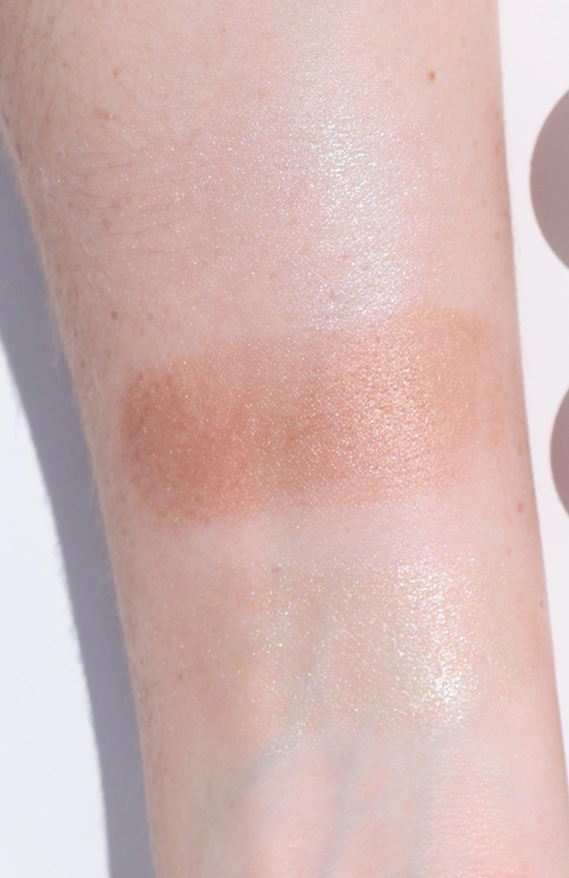 A close-up image of a forearm showing three swatches of River Organics Skincare Highlighter and Bronzer Sets. The top and bottom swatches have a shimmering, translucent finish, while the middle swatch is a metallic, peachy bronze bronzer. The skin is fair with some freckles visible.