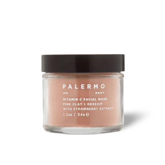 A clear glass jar with a black lid holds Palermo Body's Vitamin C Facial Mask, made with pink clay, rosehip, and strawberry extract for gentle exfoliation. The front label displays white text.