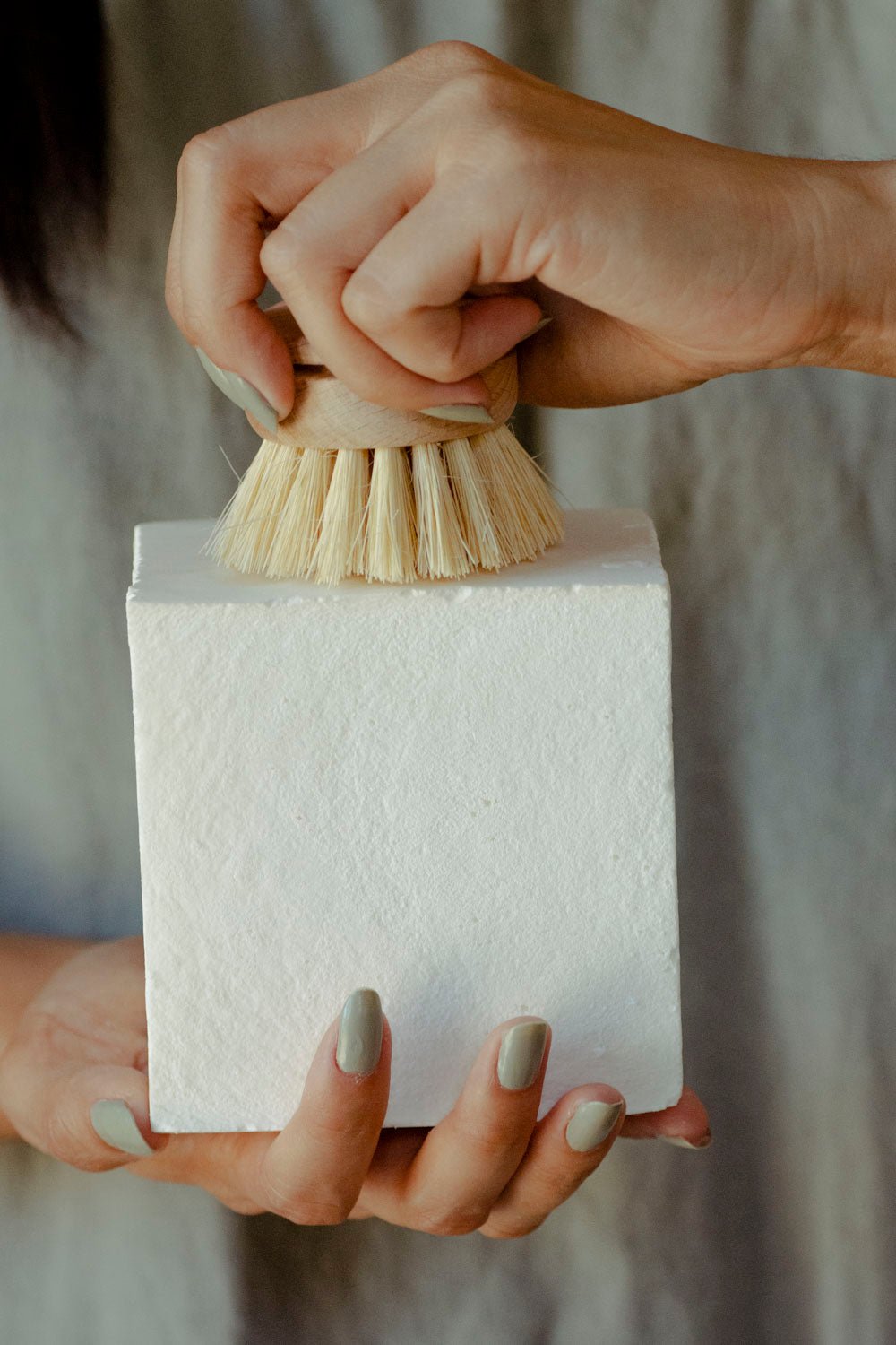 A person holds a white No Tox Life DISH BLOCK® solid dish soap (22.5 oz) in one hand and scrubs it with a wooden brush, showcasing light-colored nail polish. This huge bar is made from biodegradable vegan ingredients.