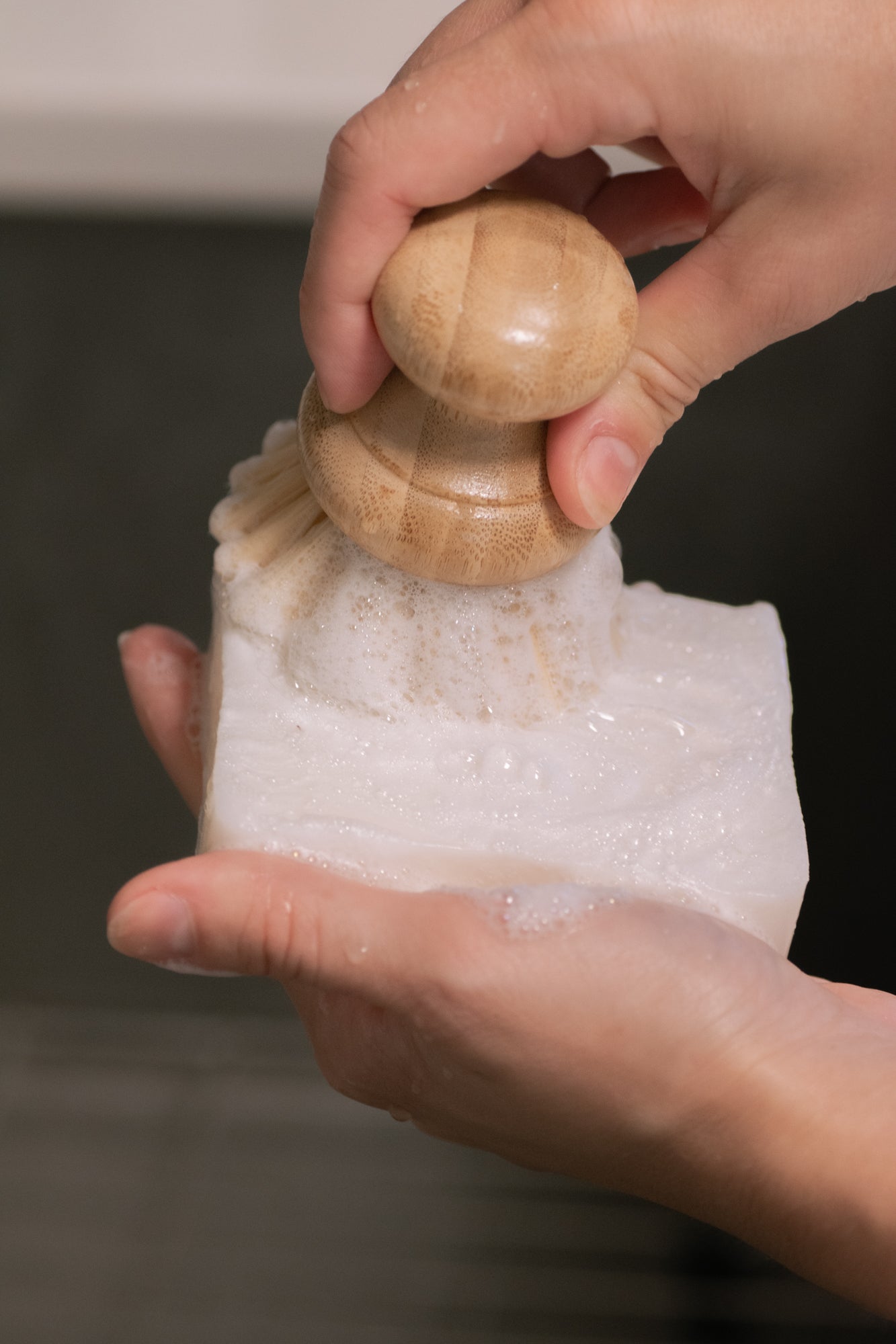 A person holds a No Tox Life DISH BLOCK® solid dish soap (6 oz) in one hand and scrubs it with a wooden brush, generating grease-cutting lather. The shot focuses on hands and the biodegradable cleaning action, with a blurred background.