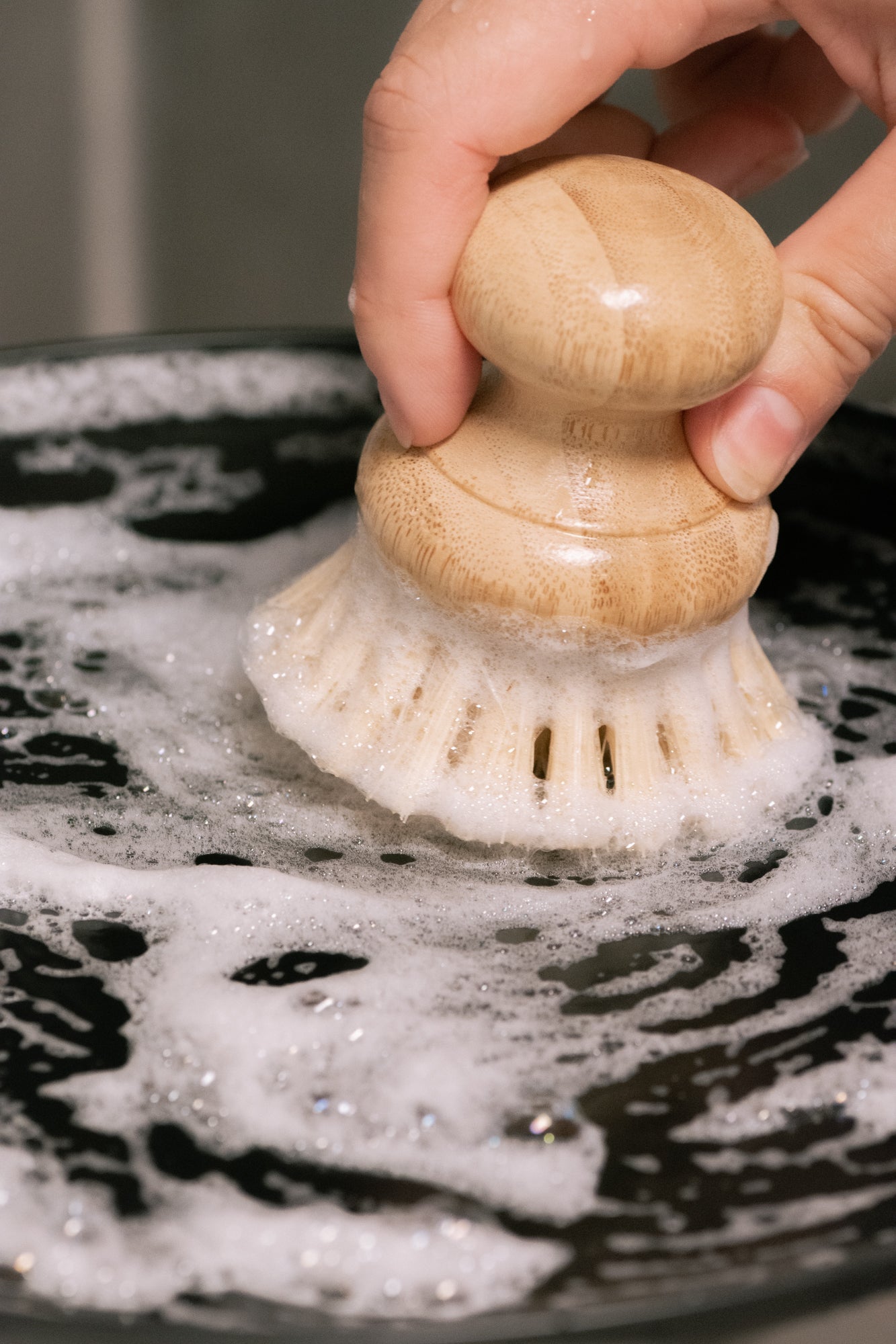 A hand scrubs a soapy, black surface with a wooden dish brush and No Tox Life DISH BLOCK® solid dish soap bar (6 oz), creating bubbles while cutting through grease.