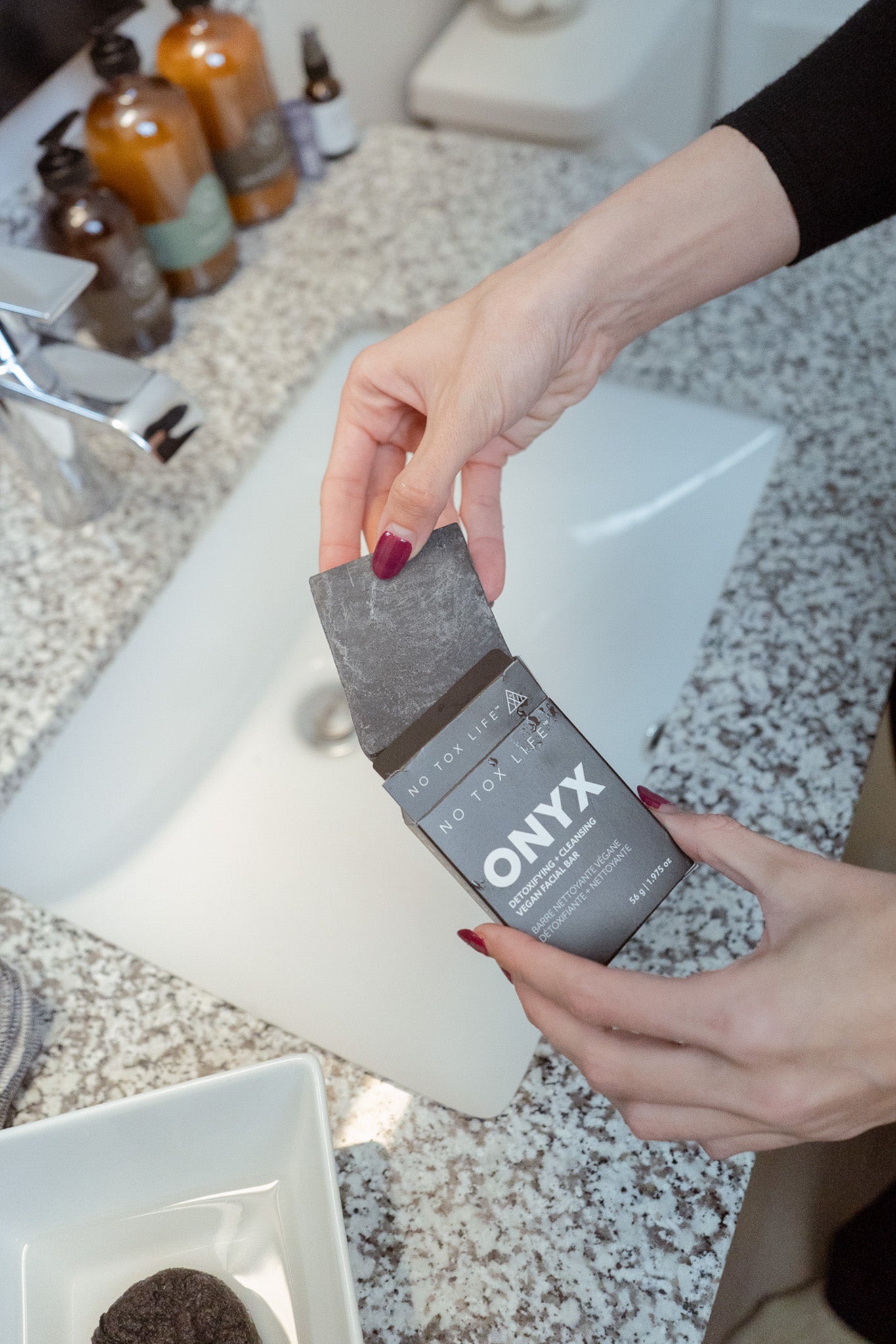 A person holds an open box of NO TOX LIFE SOLIDSILK® ONYX Facial Cleansing Bar over a granite bathroom sink, ready to remove the black bar from the packaging. Bottles and a small container sit nearby.
