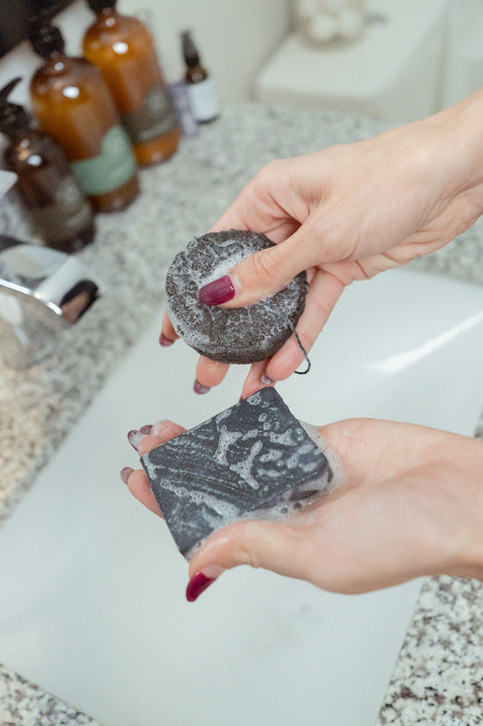 A person with maroon-painted nails holds the NO TOX LIFE SOLIDSILK® ONYX Facial Cleansing Bar and a round exfoliating sponge, both foamy, over a bathroom sink with brown bottles in the background.