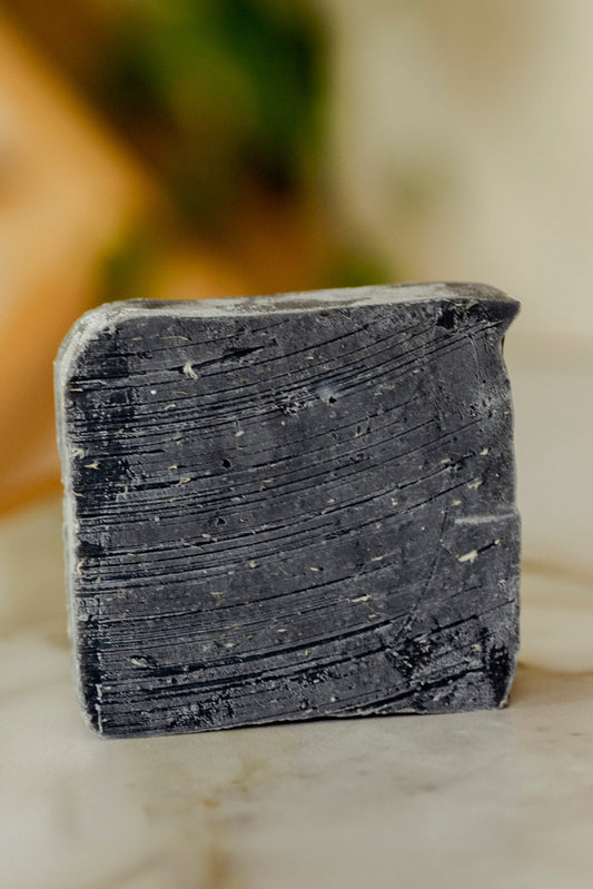 A close-up of the No Tox Life Underarm Detox Bar (large), a rectangular black soap with textured lines for exfoliation and underarm detox, sits on a light surface with a blurred background.