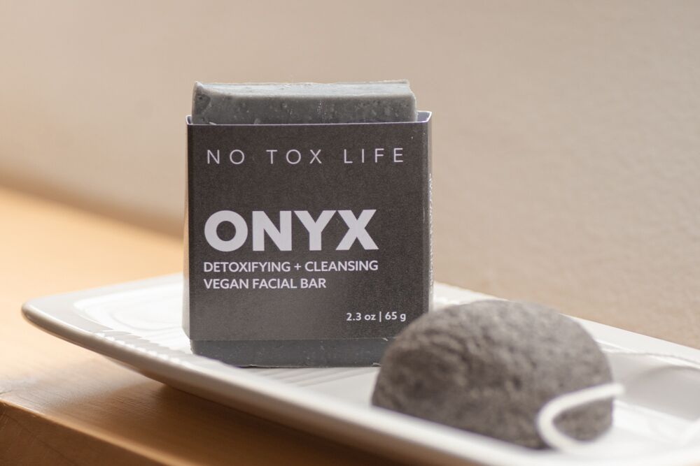 A SOLIDSILK® ONYX Facial Cleansing Bar by NO TOX LIFE sits on a white tray next to a round gray sponge, both softly lit by natural light.