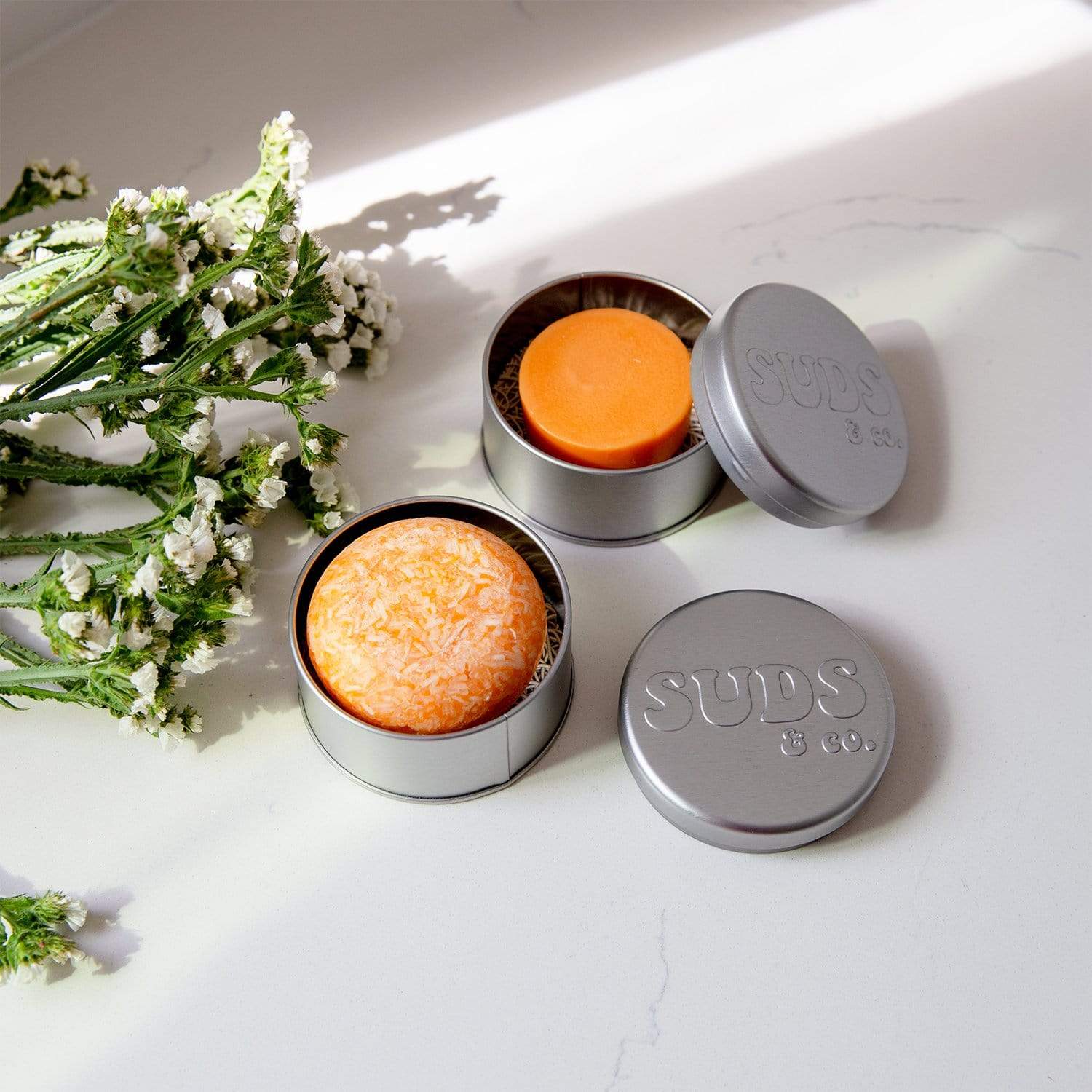 Two silver tins from ZeroWasteStore.com sit on a white surface, each holding a vibrant orange Shampoo & Conditioner Bar Duo—ideal for zero waste routines—with small white flowers nearby and soft sunlight casting shadows.