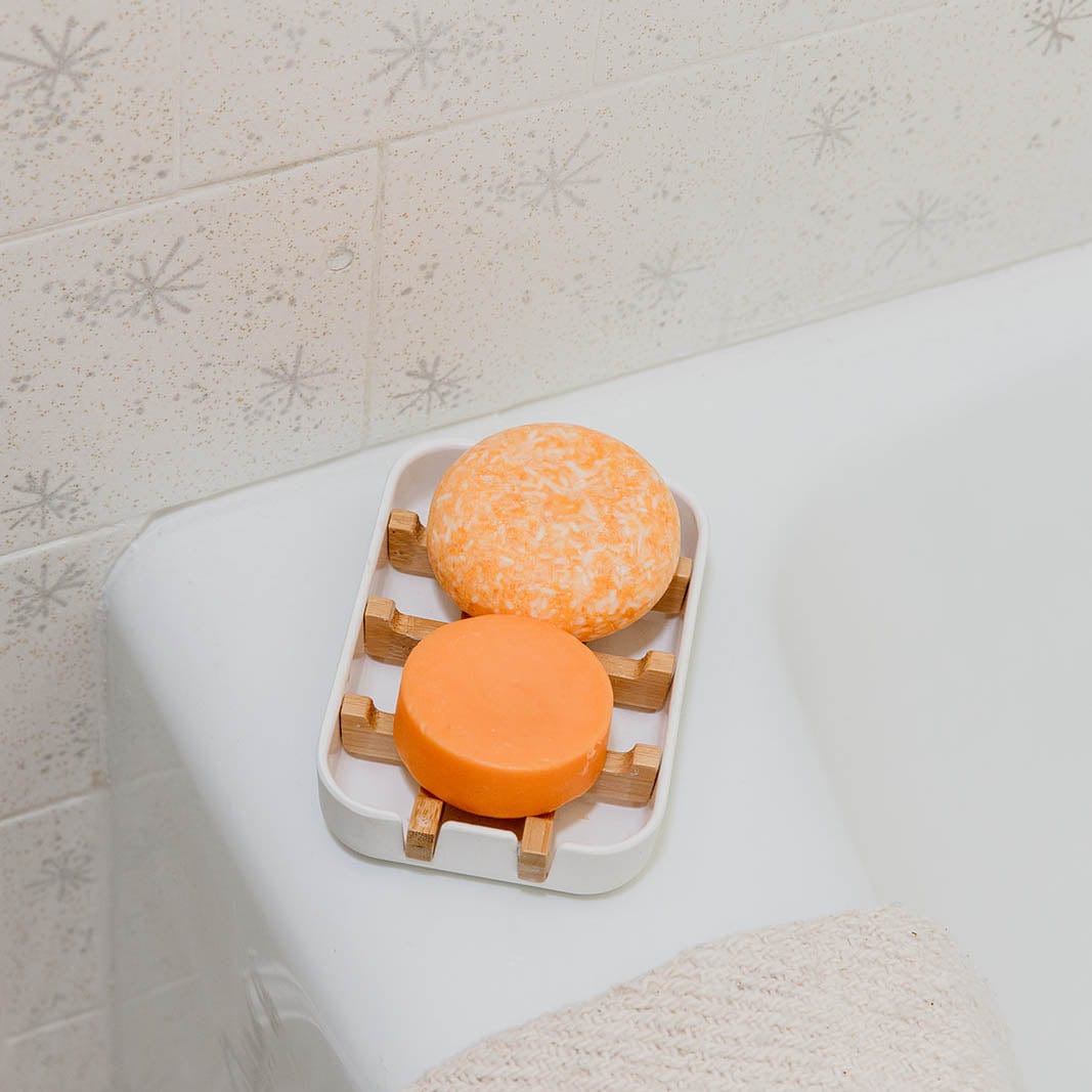 Two orange Shampoo & Conditioner Bar Duo bars from ZeroWasteStore.com sit on a soap dish by the bathtub with a towel and patterned tile wall, adding a zero waste touch to your bathroom.