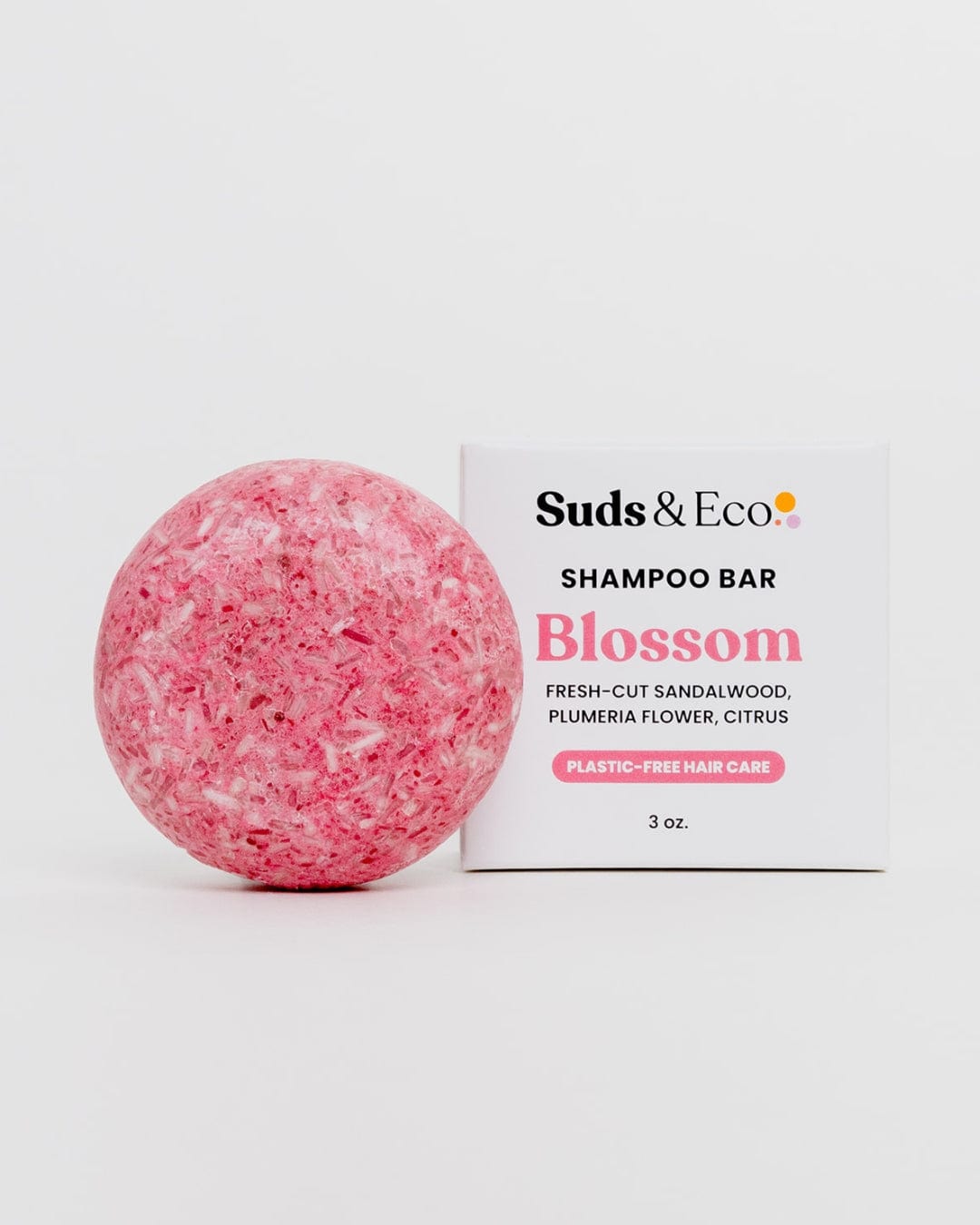 A pink round Shampoo Bar from ZeroWasteStore.com sits by its white box labeled “Shampoo Bar, Blossom, sandalwood, plumeria, citrus, vegan & SLS-free, plastic-free hair care, 3 oz.” on a plain white background.