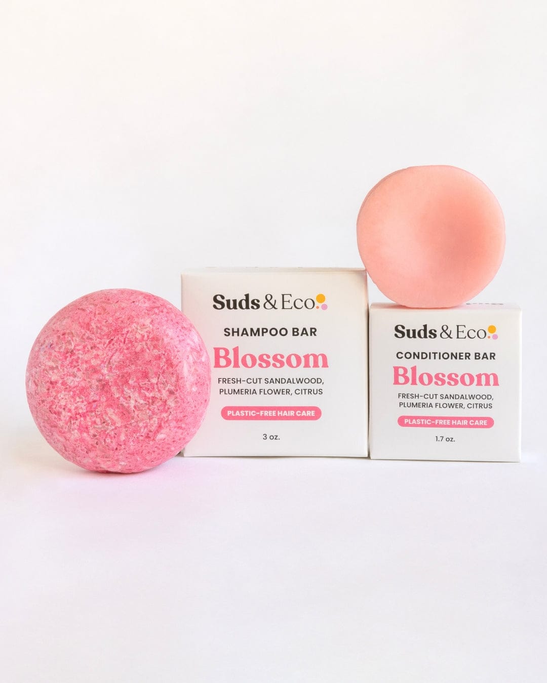 Two round, pink bars—a larger speckled one and a smaller smooth one—stand before white boxes labeled “ZeroWasteStore.com.” The Shampoo & Conditioner Bar Duo features vegan packaging and floral-citrus scents for a zero waste hair routine.