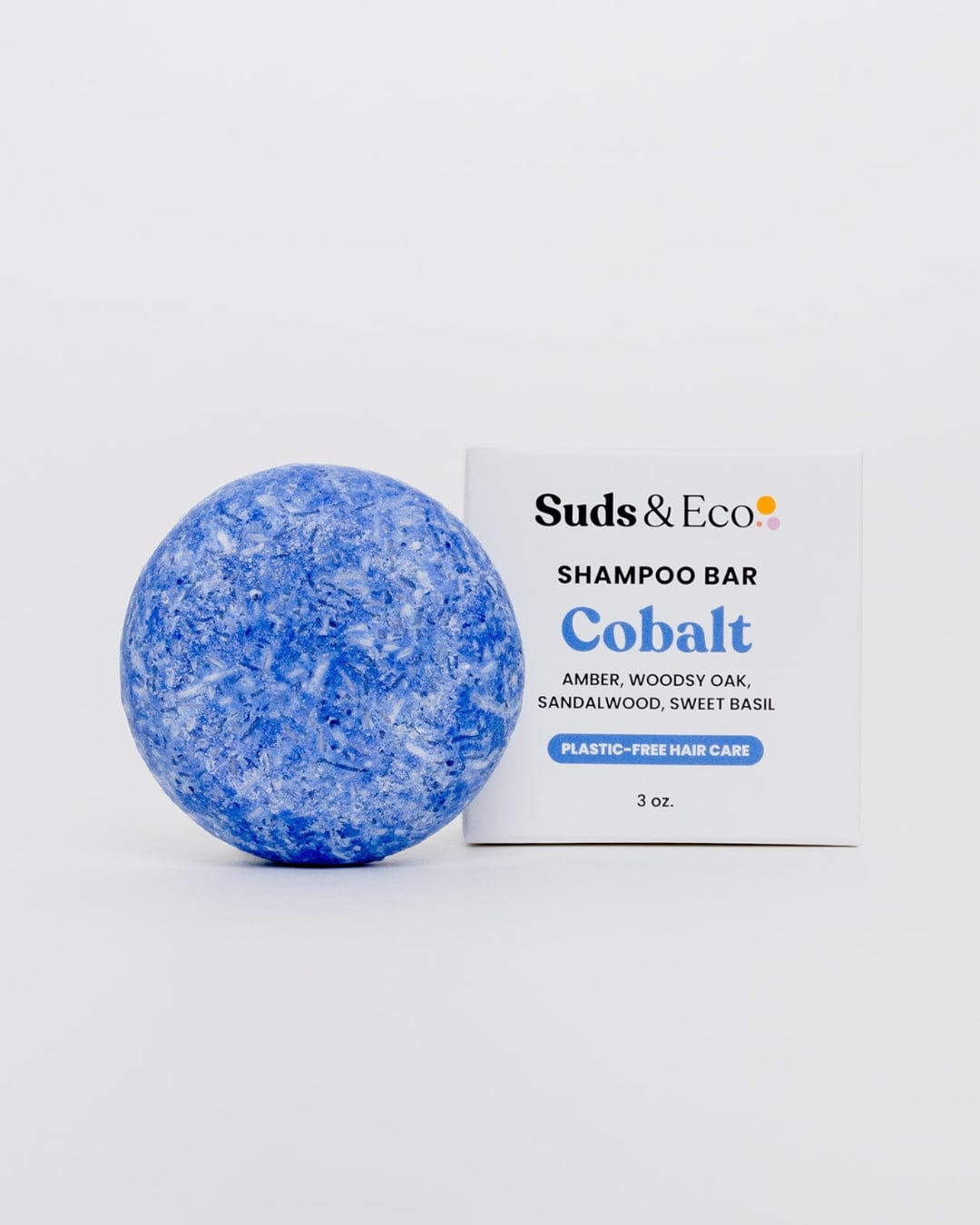 A round, blue Shampoo Bar from ZeroWasteStore.com rests by its box labeled “Shampoo Bar, Cobalt,” featuring amber, oak, sandalwood, and sweet basil scents. The packaging highlights plastic-free hair care.