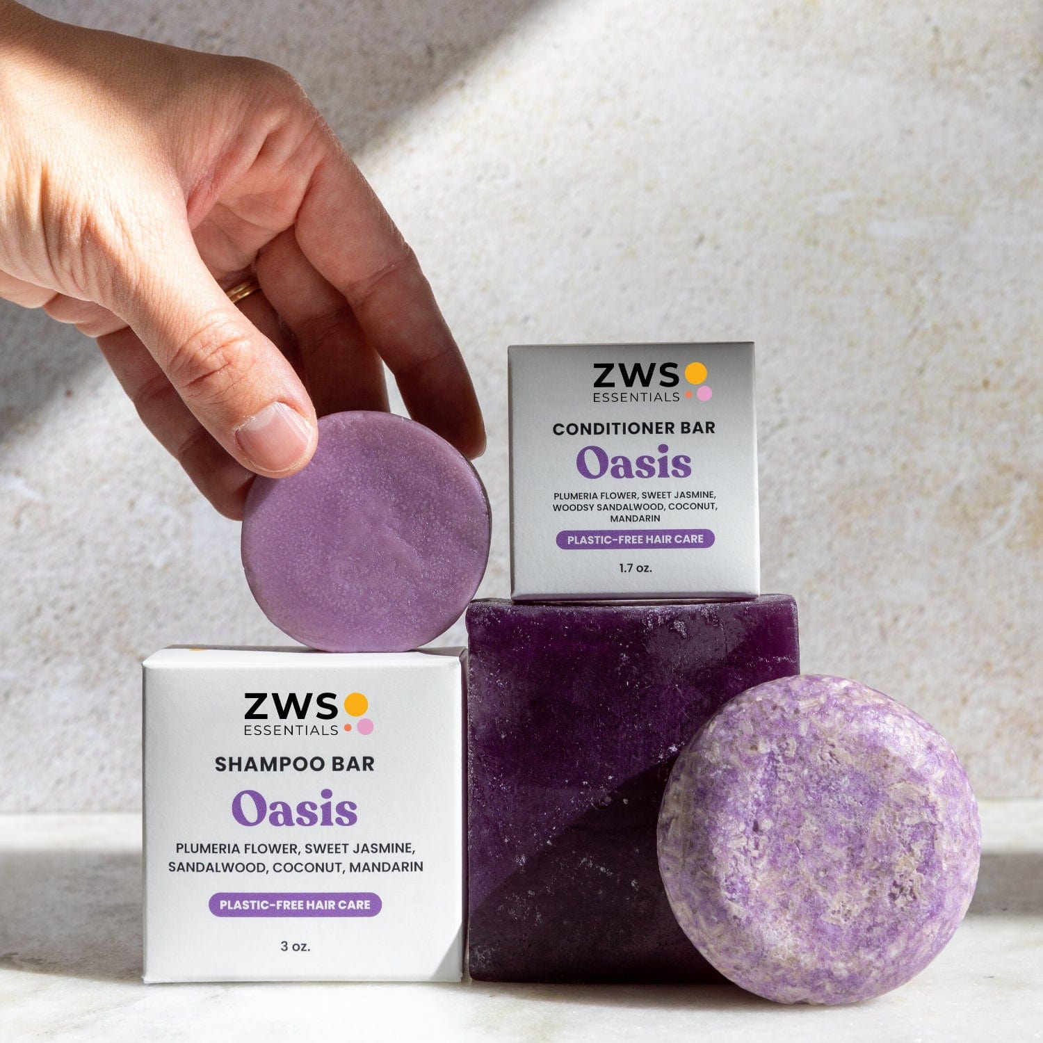 A hand holds a round purple Conditioner Bar from ZeroWasteStore.com above boxed sets, with two more bars nearby on a sunlit surface.