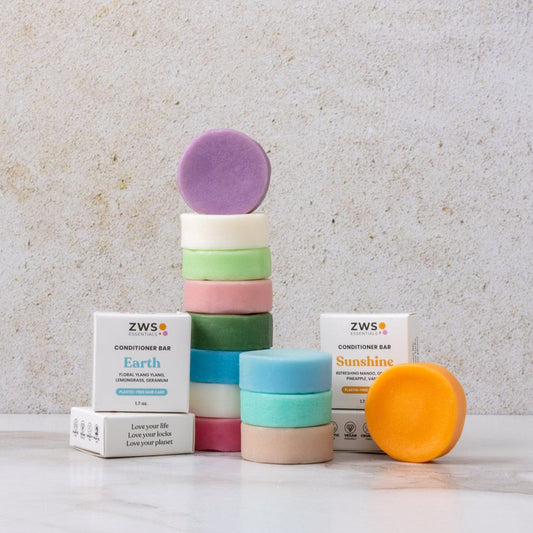 A stack of colorful ZeroWasteStore.com Conditioner Bars, including a vegan zero waste bar, is arranged between two white "Earth" and "Sunshine" boxes. A purple bar sits on top; an orange bar leans against a box, all on a light background.