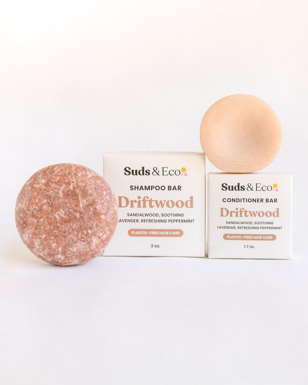 Two round bars—one pinkish-brown, one beige—sit before white ZeroWasteStore.com boxes. The Shampoo & Conditioner Bar Duo in Driftwood scent offers an eco-friendly hair care routine with zero-waste packaging.
