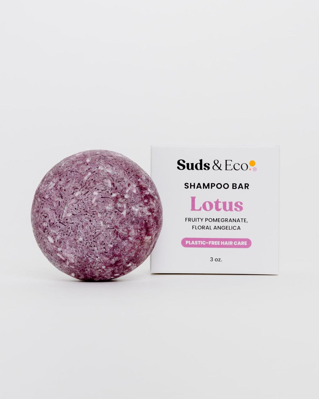 A round, purple Shampoo Bar from ZeroWasteStore.com sits next to a white box labeled "Shampoo Bar Lotus, Fruity Pomegranate, Floral Angelica, Plastic-Free Hair Care, 3 oz." on a plain white background.