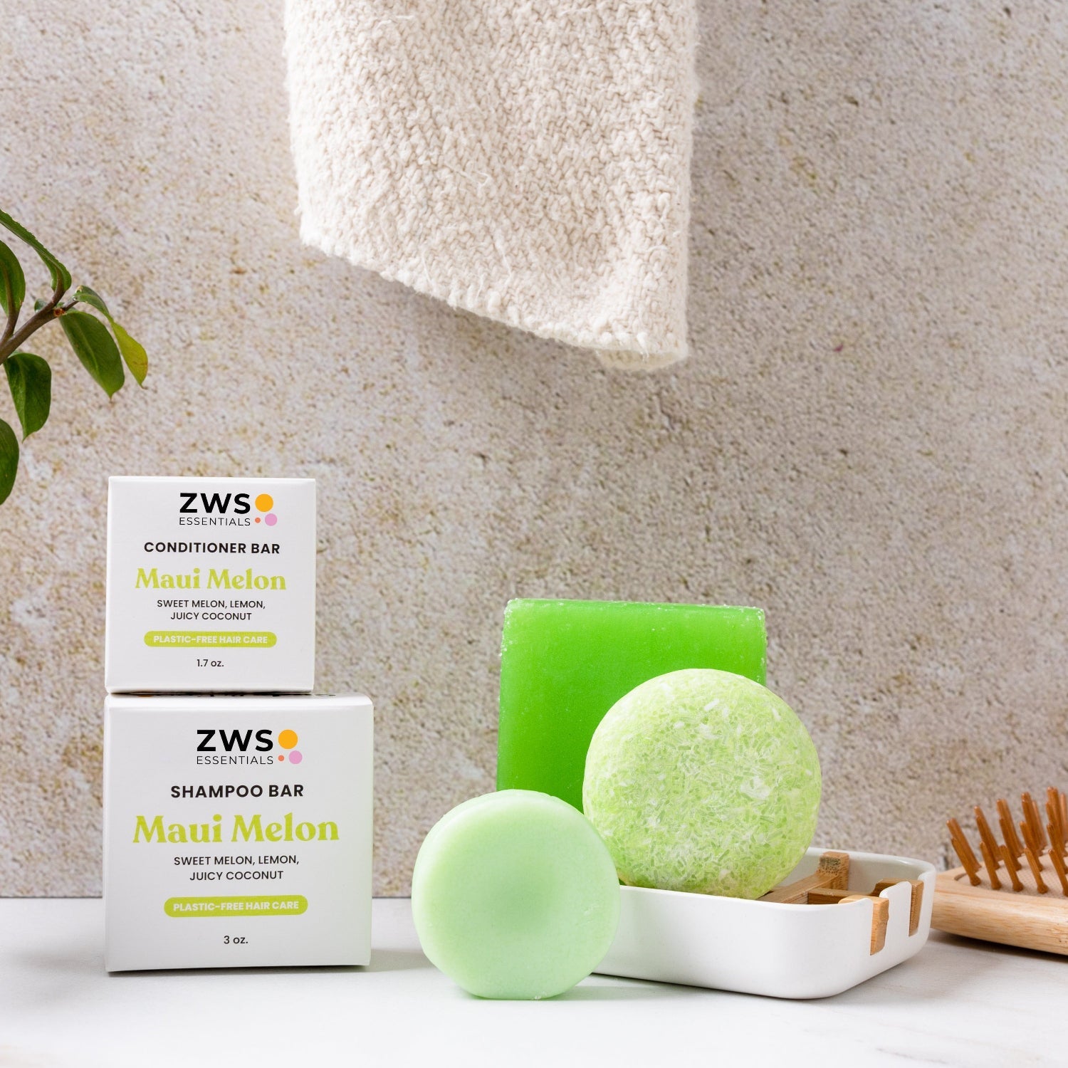 ZeroWasteStore.com's green Shampoo Bar and round conditioner bar with packaging are displayed on a white surface, alongside a wooden soap dish, green plant, and a hanging beige towel in the background.