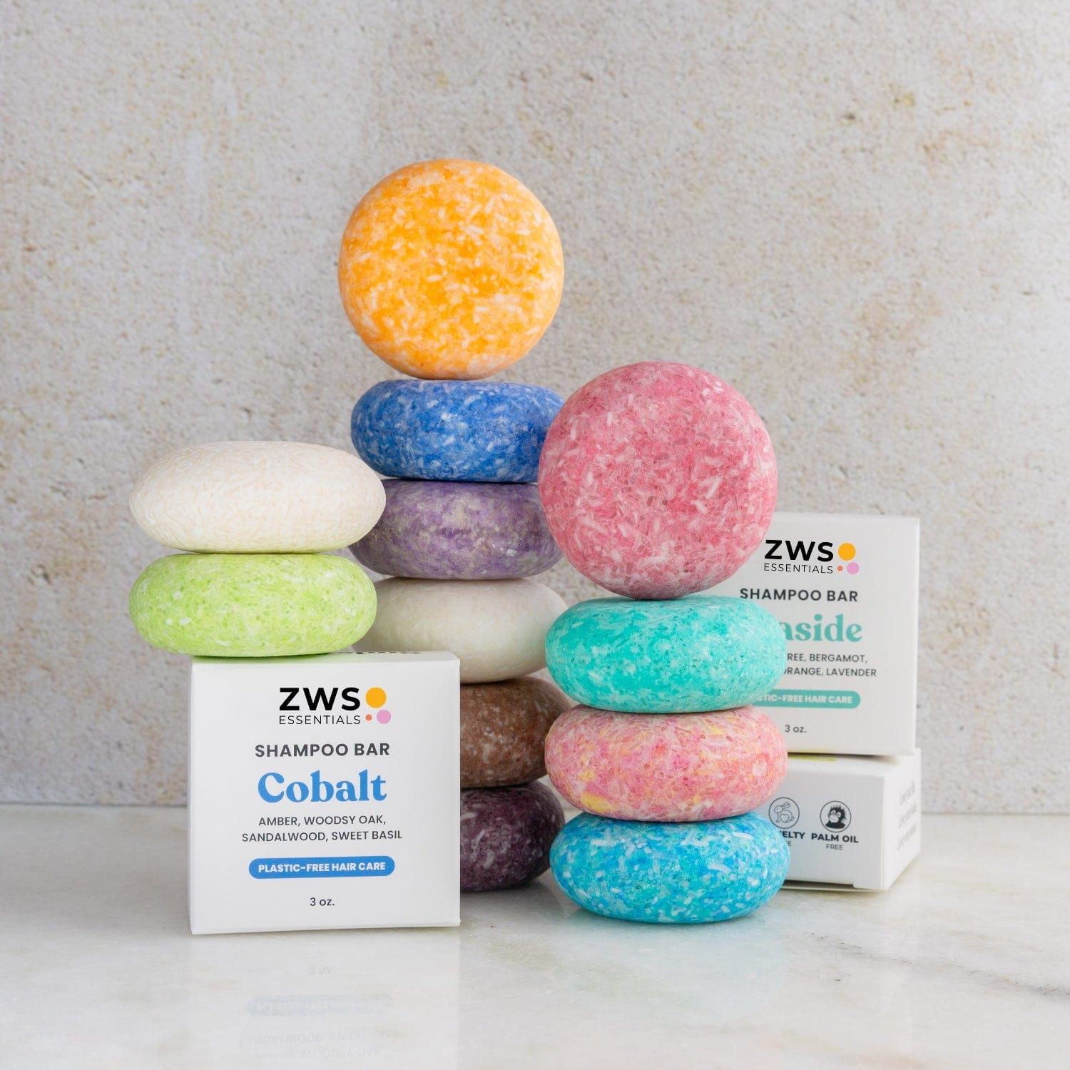A stack of colorful Shampoo Bars from ZeroWasteStore.com—yellow, purple, white, green, blue, pink, orange—sits beside ZWS Essentials "Cobalt" and "Lakeside" boxes on a neutral background.