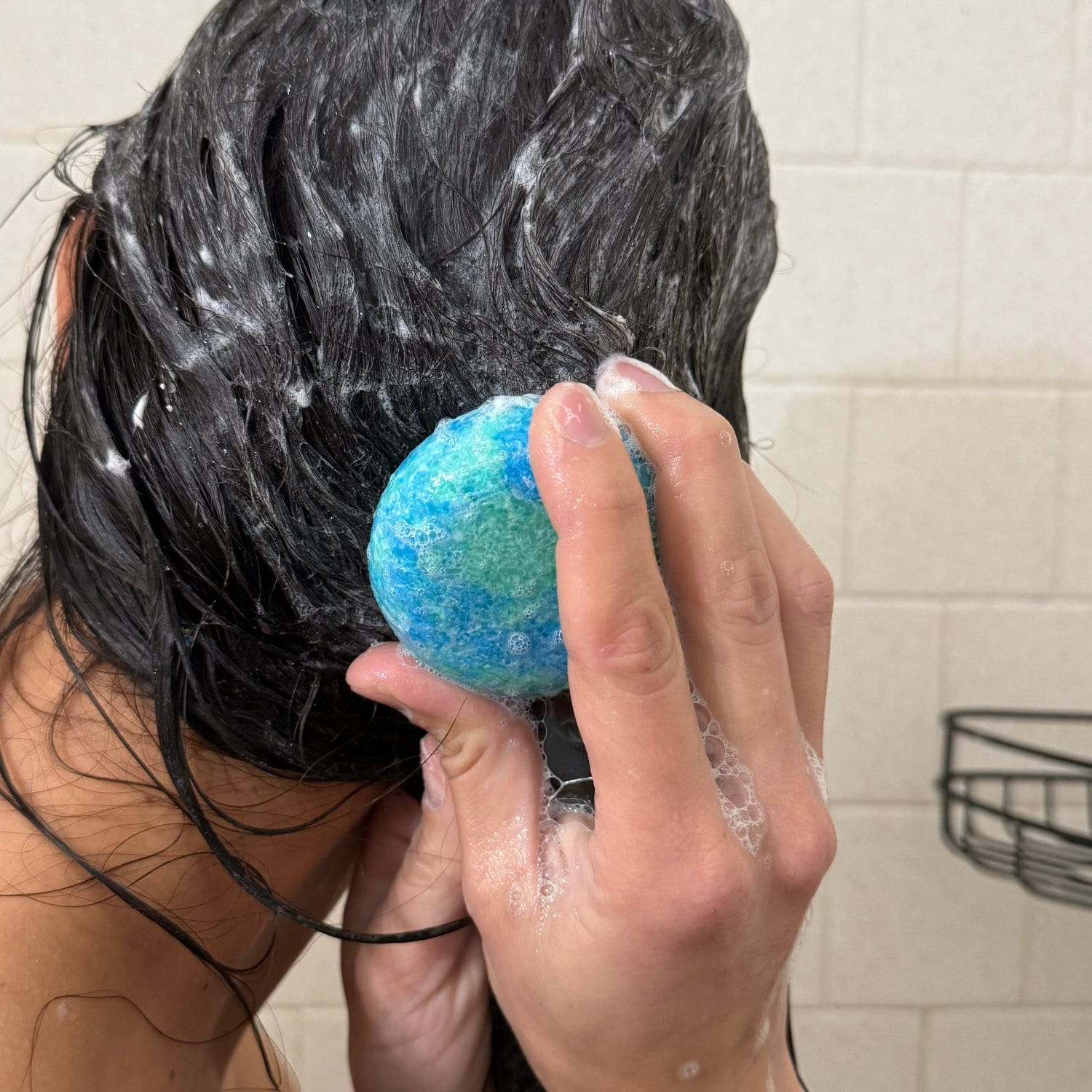 A person with wet, dark hair uses the blue and green ZeroWasteStore.com Shampoo Bar to lather their hair in a tiled shower, with visible soap suds on their hair and hand.