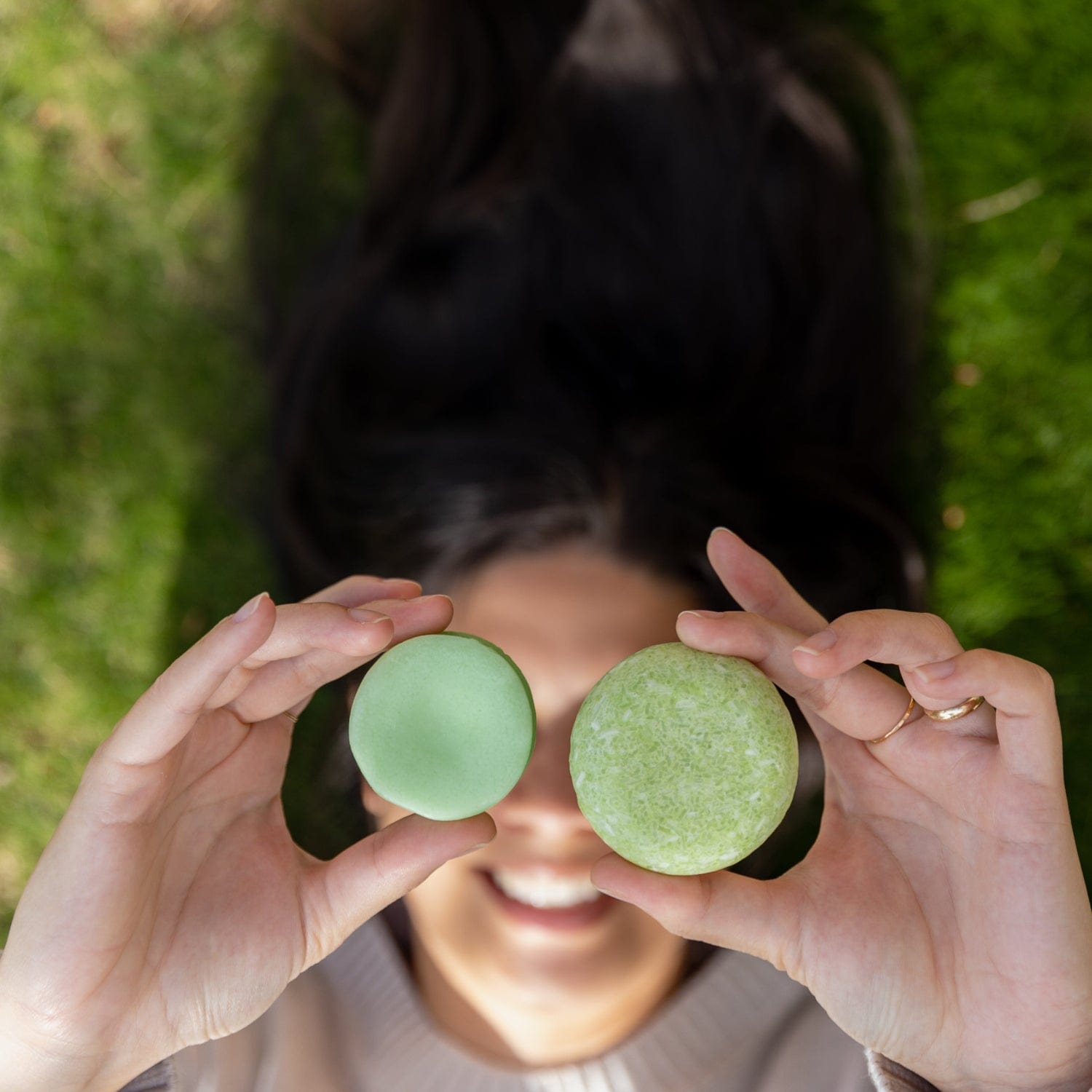 A person lying on grass smiles, holding a green macaron and round object aloft. Their long dark hair—shiny from the Shampoo Bar by ZeroWasteStore.com—spreads out behind them.