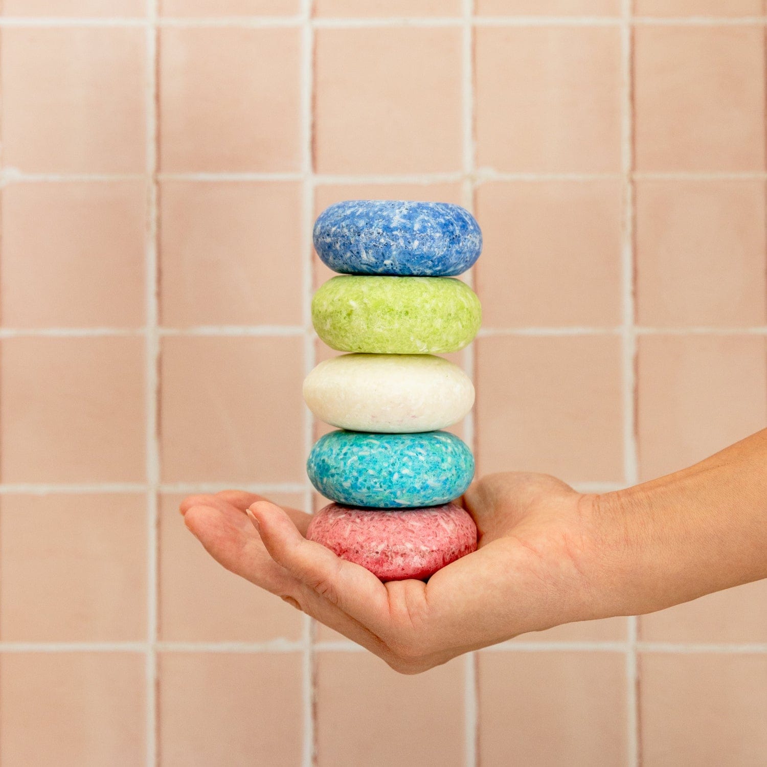 A hand holds a vertical stack of five round, colorful Shampoo Bars from ZeroWasteStore.com—blue, green, white, turquoise, and pink—in front of a light pink tiled wall.