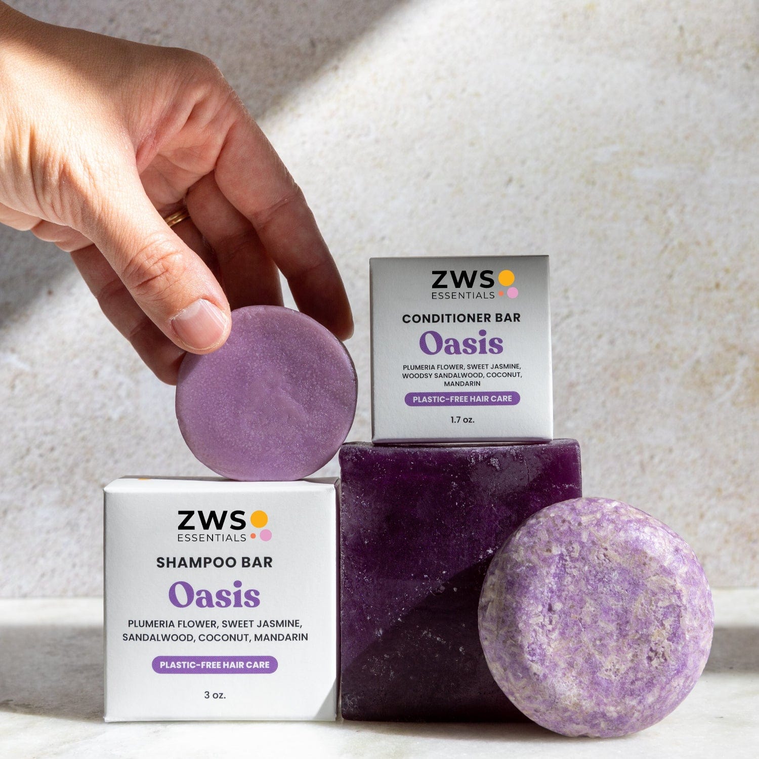 A hand holds a round purple Shampoo & Conditioner Bar Duo by ZeroWasteStore.com next to two branded boxes, with another purple bar and sunlight casting eco-friendly shadows in the background.