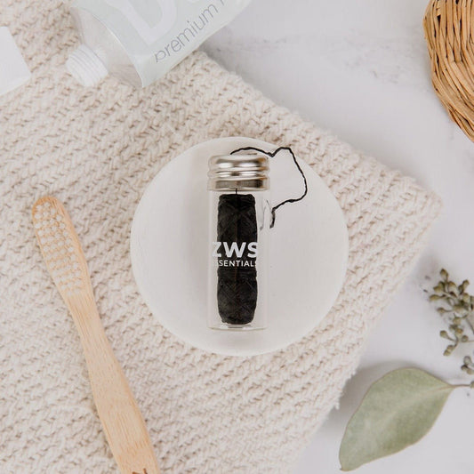 A small glass jar of Bamboo Charcoal Floss by ZeroWasteStore.com rests on a white dish, alongside a bamboo toothbrush, toothpaste tube, woven basket, and leafy sprigs atop a textured mat.