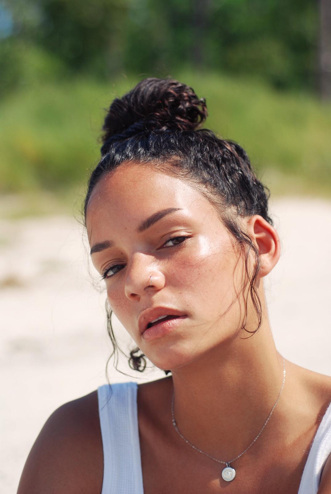 A person with curly hair tied in a top knot bun gazes directly at the camera. They are wearing a white sleeveless top and a necklace with a circular pendant. The background is blurred, showing a bright, outdoor environment with greenery, subtly illuminated by River Organics Skincare Vegan Highlighter Stick | Rose Quartz.