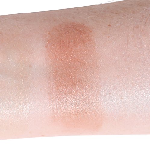 A close-up of a forearm shows a swatch from River Organics' Shimmery Face and Body Bronzer Stick, featuring vegan ingredients. The reddish-brown bronzer contrasts beautifully with the surrounding skin.