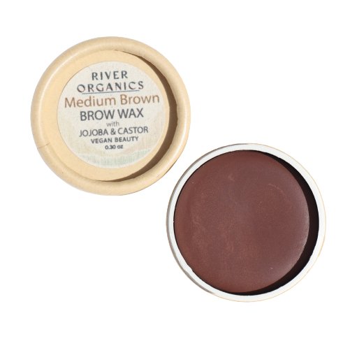 A small container of River Organics Skincare Tinted Vegan Eyebrow Wax | Medium Brown is shown with its lid removed. The lid, placed next to the container, displays the product label indicating that this tinted eyebrow wax is made with jojoba and castor and is both vegan and cruelty-free certified. The brow wax inside is a rich brown color.