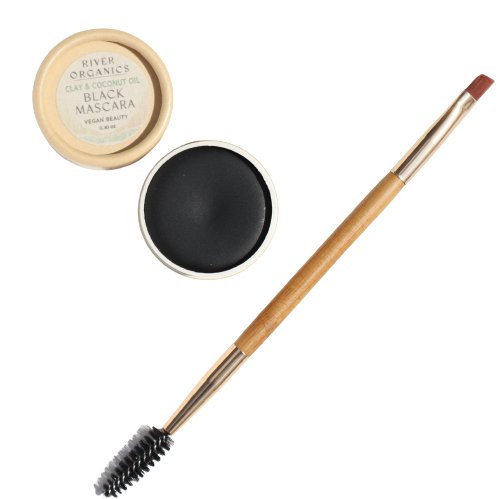 An open container of River Organics Black Zero-Waste Mascara, made with clay and coconut oil, is placed beside a wooden makeup brush with a spoolie on one end and an angled brush on the other. This chemical-free mascara is labeled as vegan beauty.