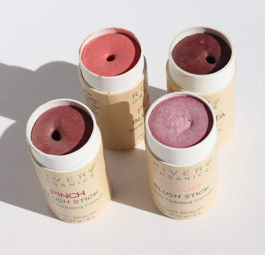 Four River Organics Imperfect Creamy Vegan Blush Sticks, with open caps displaying cream-to-powder pink and mauve shades, are arranged on a bright white surface. The eco-friendly cylindrical packaging shows their shade names. Available in limited quantities.