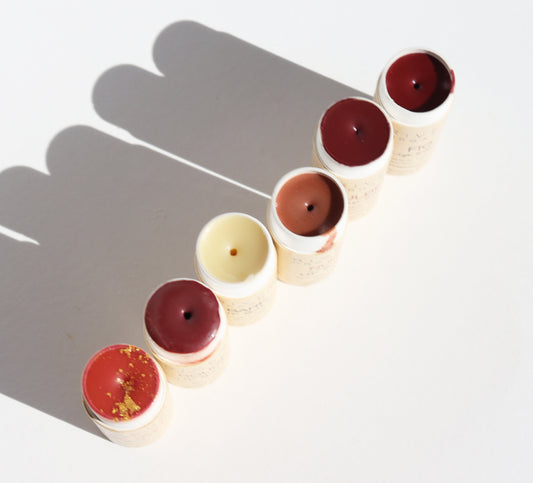 Six full-size Imperfect Lip Balms + Lip Stains from River Organics are diagonally arranged on a white surface, with opened tubes showcasing shades of red, pink, brown, and cream. Shadows add texture to the left of the display.