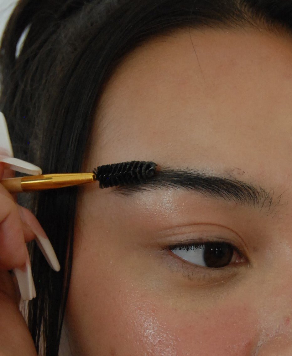 A close-up of a person grooming their eyebrow with a spoolie brush, using River Organics Tinted Vegan Eyebrow Wax | Dark. The hand holding the brush has white nails. The eyebrow is dark and thick, and the person's hair is dark brown. Only part of the person's face from the eye to the forehead is visible.